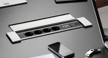 In einen Konferenztisch integrierte Steckdosenleiste mit USB-Anschlüssen, LAN-Ports und mehreren Geräten auf dem Tisch.
